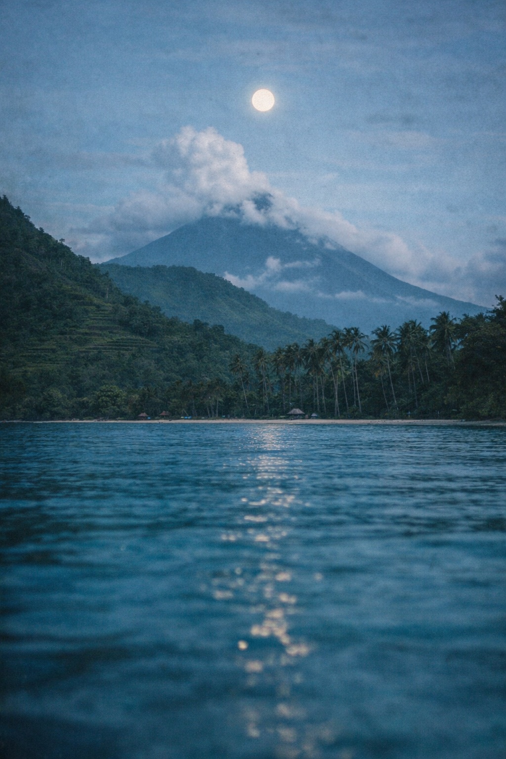 A full moon over a clouded volcano at twilight, its light running down the water to a palm-lined shore.